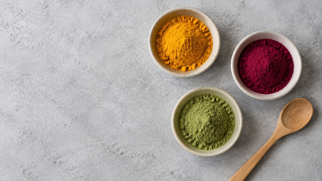Illustrative bowls of golden, magenta and green botanical powders on a gray concrete surface.