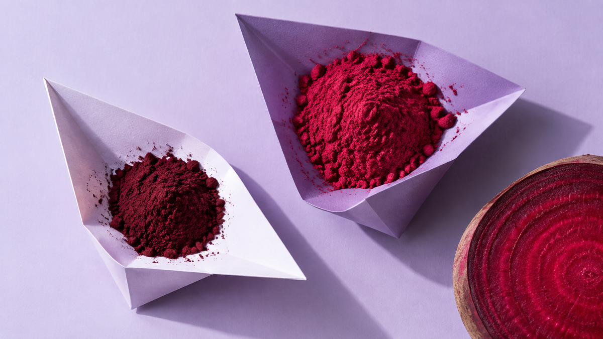 Two folded paper scoops holding separate burgundy and beet-red powder mounds on a pale lavender background beside a cropped beet slice